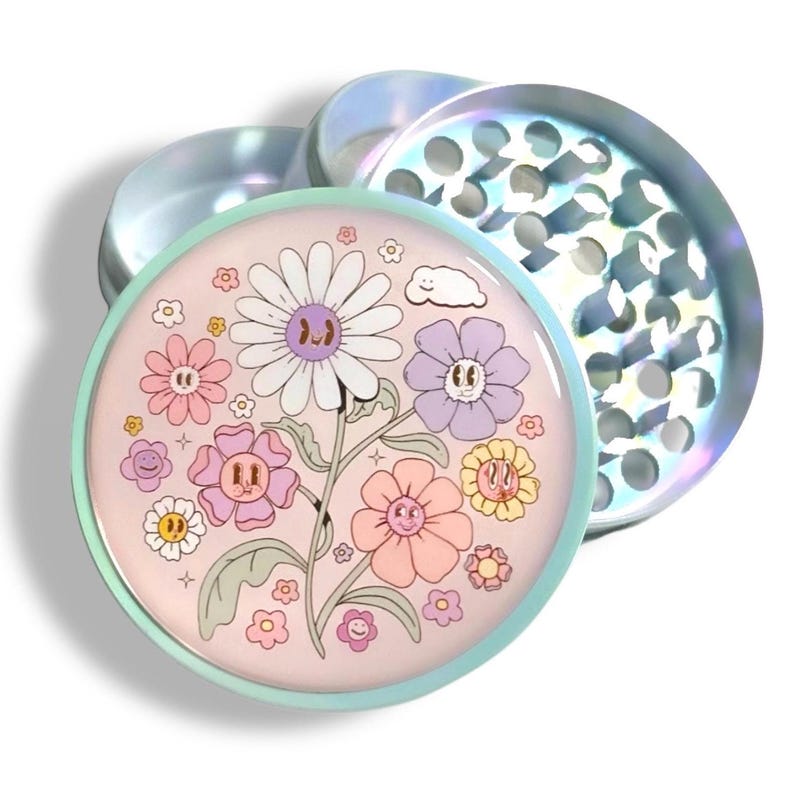 May include: A two-piece, iridescent grinder with a floral design. The top piece is a lid with a series of holes. The bottom piece has a light pink background with a variety of cartoon flowers in shades of pink, purple, yellow, and white.