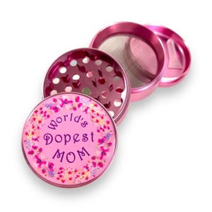 Puede incluir: Un molinillo de hierbas rosa, desmontado parcialmente, con el texto "World's Dopest MOM" y un diseño floral. El molinillo tiene una pantalla de malla y un pequeño raspador. El molinillo es de metal y tiene múltiples compartimentos.