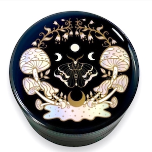 Pode incluir: Um moedor redondo preto com um design celestial. O design apresenta uma mariposa, cogumelos, luas e estrelas em cores douradas e iridescentes. O moedor &eacute; feito de metal.