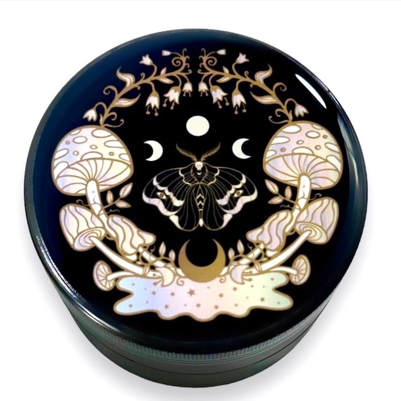 Pode incluir: Um moedor redondo preto com um design celestial. O design apresenta uma mariposa, cogumelos, luas e estrelas em cores douradas e iridescentes. O moedor &eacute; feito de metal.