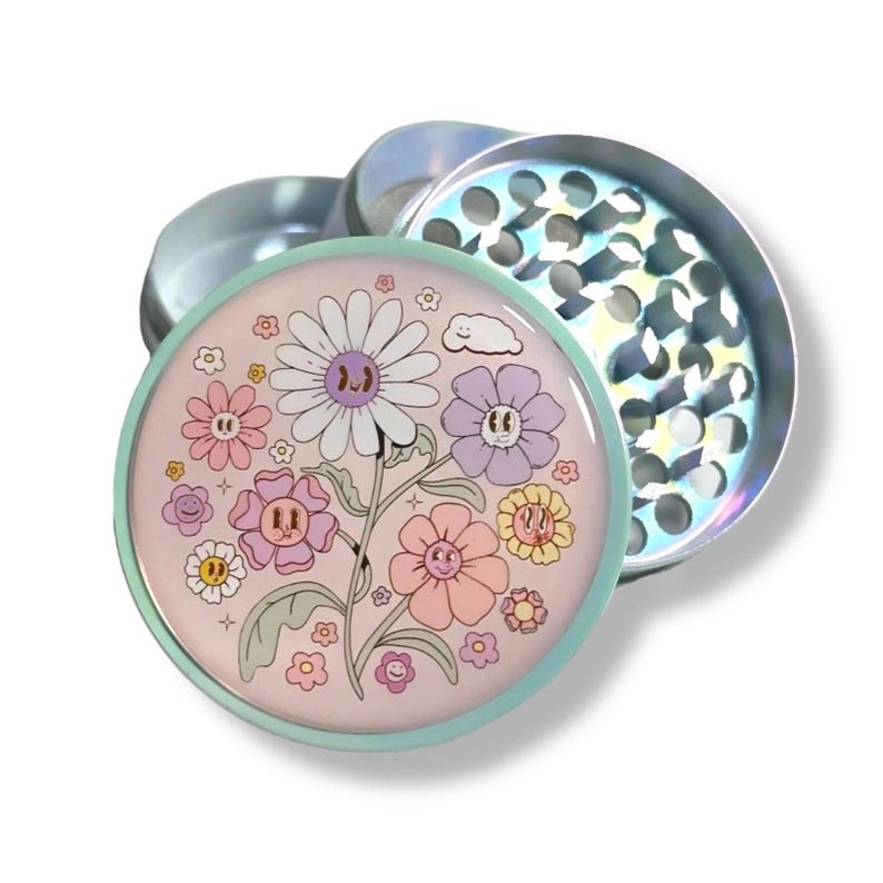 May include: A round, silver herb grinder with a pastel pink lid featuring a floral design with smiling flowers and a cloud. The grinder has a light blue rim and a silver, perforated grinding chamber.