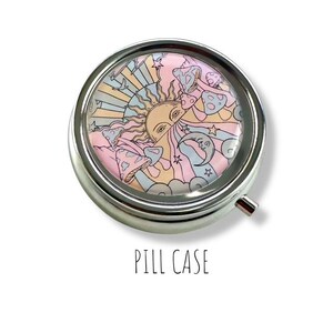 Peut inclure: Un pilulier rond et argent&eacute; avec un motif psych&eacute;d&eacute;lique color&eacute;. Le motif repr&eacute;sente un soleil, des champignons, des &eacute;toiles et un croissant de lune aux couleurs pastel. Le texte "PILL CASE" est en dessous.