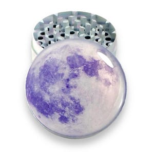 May include: A silver metal grinder with a purple and white moon design on the lid. The grinder has a circular shape and a textured surface.