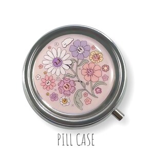 May include: A circular, silver pill case with a floral pattern. The design shows smiling flowers in pink, purple, and yellow on a pale pink backdrop. The text "PILL CASE" is printed in black beneath the case.