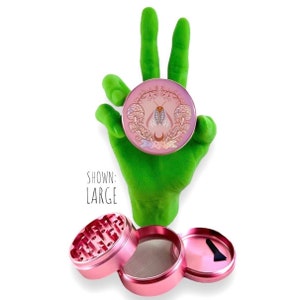 May include: A pink metal grinder with a design of a moth and mushrooms. The grinder has four parts, including a lid, a top chamber, a screen, and a bottom chamber. The text "SHOWN: LARGE" is visible in the image.