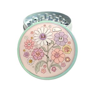 May include: A round, silver metal herb grinder with a floral design on the lid. The lid features a light pink background with smiling flowers in various colors, including white, purple, and peach. The grinder has a light blue rim.