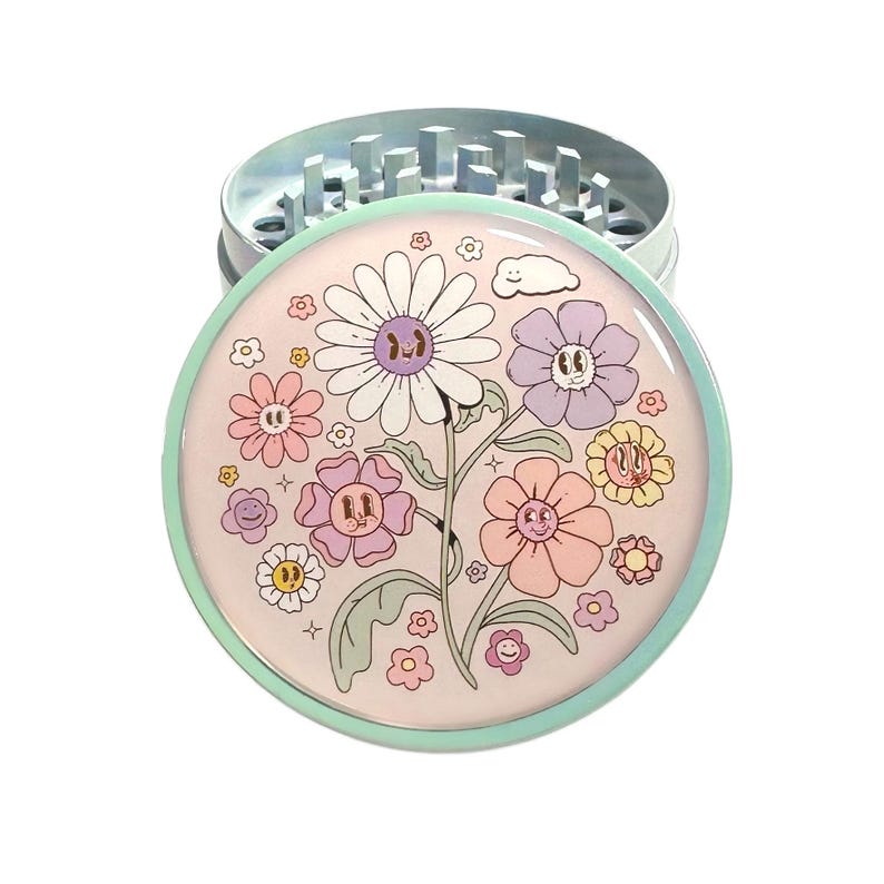 May include: A round, silver metal herb grinder with a floral design on the lid. The lid features a light pink background with smiling flowers in various colors, including white, purple, and peach. The grinder has a light blue rim.