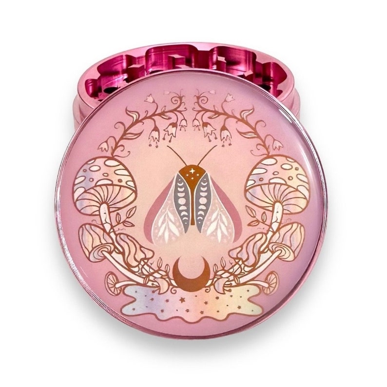 May include: A pink metal grinder with a circular design featuring a moth with a crescent moon, mushrooms, and floral accents.