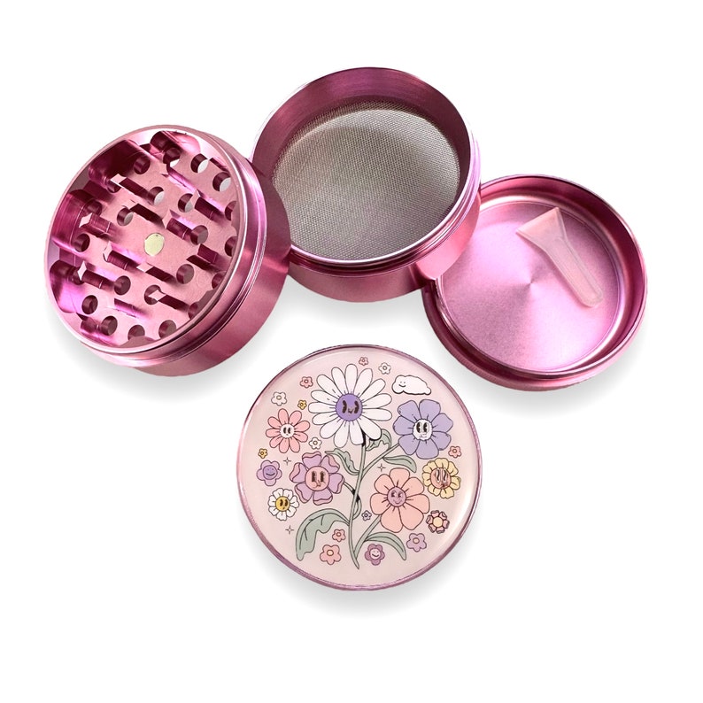 May include: A disassembled pink metal herb grinder with a floral design on the lid. The grinder is composed of multiple parts, including a grinding chamber, a mesh screen, and a storage compartment. A small plastic scraper is included.