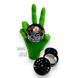 Puede incluir: Un grinder de hierbas negro con un dise&ntilde;o floral y de serpiente. El grinder se muestra con una mano verde haciendo el signo de la paz. El texto "SHOWN: LARGE" es visible. El grinder es negro con una malla plateada.