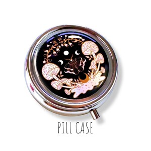 Pode incluir: Um estojo de comprimidos redondo e prateado com fundo preto. O estojo apresenta um desenho de cogumelos, uma tra&ccedil;a e luas crescentes. O texto "PILL CASE" est&aacute; impresso em preto na parte inferior da imagem.