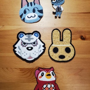 May include: Five embroidered patches featuring characters from the Animal Crossing video game series. The patches are on a wooden surface. The characters are a blue tabby cat, a white tiger, a brown bunny, a grey cat wearing glasses, and a red owl.