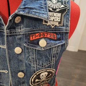 May include: A blue denim vest with several patches sewn on. The patches include a skull and crossbones, a patch with the text "Northern Capers Flight", and a patch with the text "TI-95728".