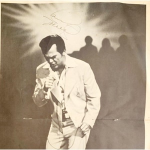 May include: Black and white photograph of a man in a light-colored suit, holding a microphone and singing. The image has a signature in the upper left corner and shadows of people in the background.