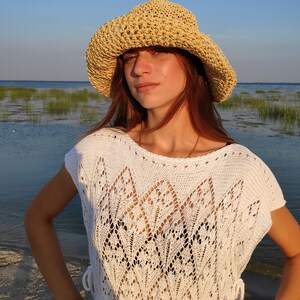 Puede incluir: Una mujer con un top de crochet blanco y un sombrero de paja. Est&aacute; de pie en una playa con agua y hierba en el fondo.