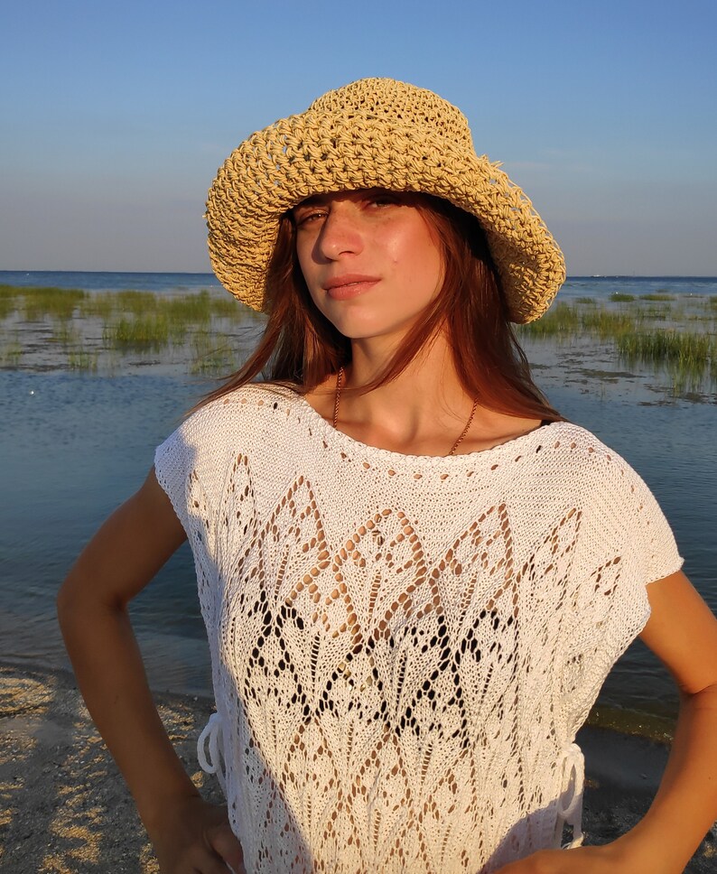 Puede incluir: Una mujer con un top de crochet blanco y un sombrero de paja. Est&aacute; de pie en una playa con agua y hierba en el fondo.