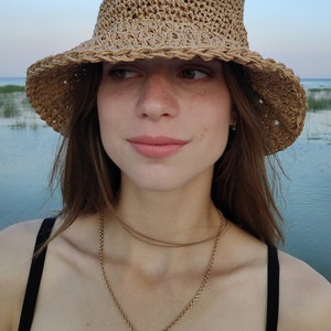 Puede incluir: Una mujer con un sombrero de paja de ala ancha y un collar de cadena dorada con un colgante de cruz negra. Est&aacute; de pie frente a una masa de agua.