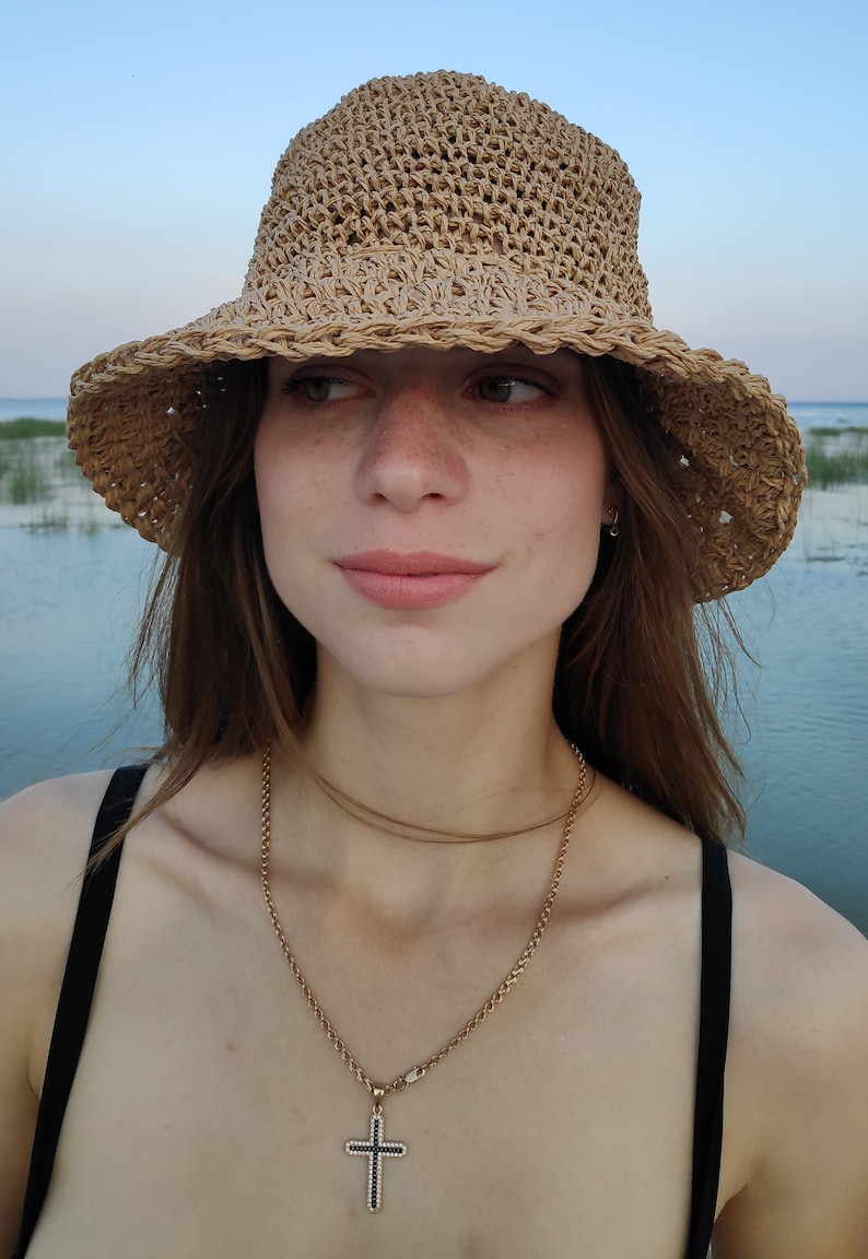Puede incluir: Una mujer con un sombrero de paja de ala ancha y un collar de cadena dorada con un colgante de cruz negra. Est&aacute; de pie frente a una masa de agua.