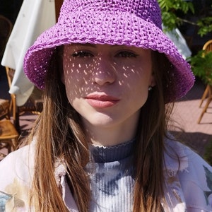 Puede incluir: Una mujer con un sombrero de sol de crochet morado de ala ancha. Lleva una chaqueta rosa claro sobre una camisa azul y blanca de tie-dye.