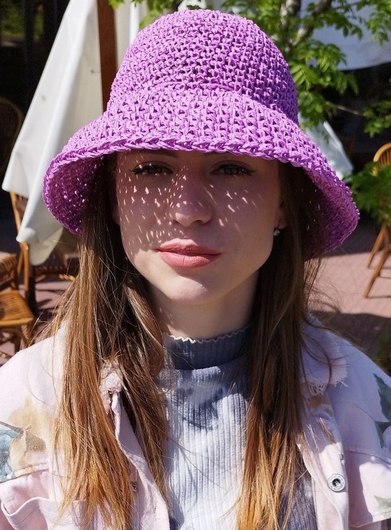 Puede incluir: Una mujer con un sombrero de sol de crochet morado de ala ancha. Lleva una chaqueta rosa claro sobre una camisa azul y blanca de tie-dye.
