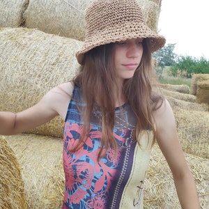 Puede incluir: Una mujer con un sombrero de paja con un cintur&oacute;n de cuero marr&oacute;n sobre un vestido sin mangas de estampado colorido. Est&aacute; de pie frente a una pila de pacas de paja.