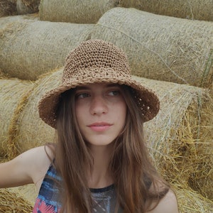 Puede incluir: Una mujer con el pelo largo y casta&ntilde;o lleva un sombrero de sol de crochet marr&oacute;n y un top de tirantes de colores. Est&aacute; de pie frente a una pila de pacas de paja.