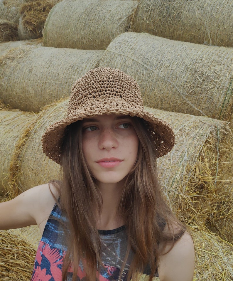 Puede incluir: Una mujer con el pelo largo y casta&ntilde;o lleva un sombrero de sol de crochet marr&oacute;n y un top de tirantes de colores. Est&aacute; de pie frente a una pila de pacas de paja.