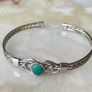 May include: A silver-toned bangle bracelet with a woven design and a turquoise-colored stone in the center. The bracelet has a hinged opening for easy wear and is displayed on a light-colored surface.