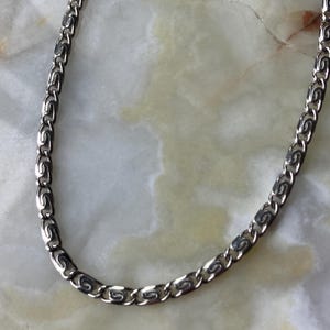 May include: A silver-colored chain necklace with a repeating pattern of interlocking links. The links have a stylized, curved design. The necklace is displayed on a light-colored, marbled surface.
