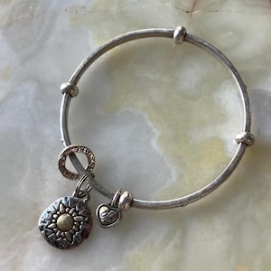 May include: Silver bangle bracelet with three decorative beads and two charms. One charm is a sunburst design with gold accents, and the other is a heart with a letter "B". The bracelet has a circular design.
