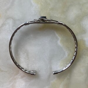 May include: Silver-toned cuff bracelet with a dark blue gemstone at the center. The bracelet features a decorative design around the gemstone and a textured, twisted rope pattern along the band. The bracelet is open at the ends.