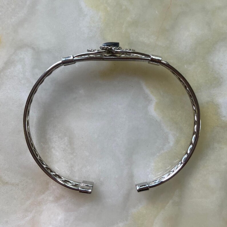 May include: Silver-toned cuff bracelet with a dark blue gemstone at the center. The bracelet features a decorative design around the gemstone and a textured, twisted rope pattern along the band. The bracelet is open at the ends.