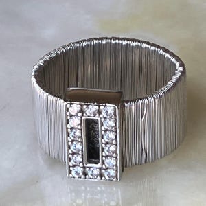 May include: A silver-toned ring featuring a wide band constructed from multiple thin, parallel metallic wires. A rectangular centerpiece is attached to the front, bordered by small, clear, sparkling stones surrounding a hollow center.