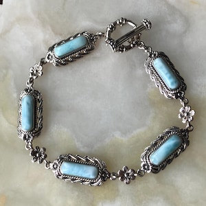 May include: A silver-toned bracelet featuring four rectangular light blue stones set in ornate metal frames. The links are connected by small flower-shaped metal components, and the piece is secured with a circular toggle clasp closure.