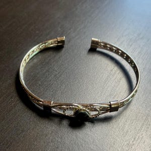 May include: Silver-toned cuff bracelet with an open design. The bracelet features a woven pattern and a small, dark-colored stone at the center. The bracelet is approximately 2.5 inches in diameter.