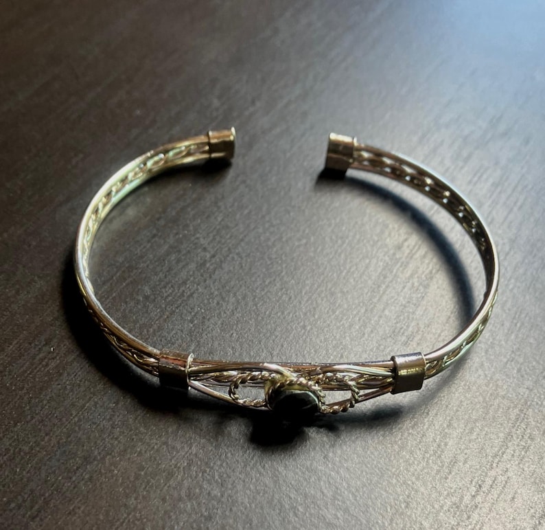 May include: Silver-toned cuff bracelet with an open design. The bracelet features a woven pattern and a small, dark-colored stone at the center. The bracelet is approximately 2.5 inches in diameter.