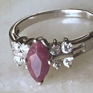 May include: A silver ring featuring a marquise-cut ruby gemstone as the centerpiece. The ring is accented with small, round, clear gemstones on either side of the ruby. The band is a simple, polished silver.