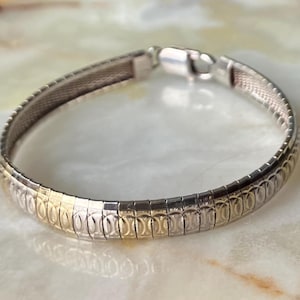 May include: A silver and gold-tone bracelet with a textured, oval-link design. The bracelet features a secure clasp and is displayed on a light-colored surface. The jewelry piece is a classic accessory.