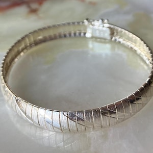 May include: A silver-colored bracelet with a textured, woven design. The bracelet has a clasp closure and a flexible, ribbed construction. The jewelry is displayed on a light-colored surface.