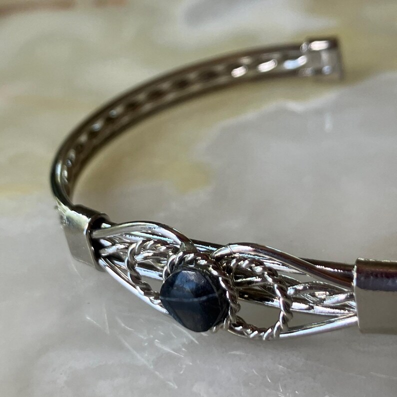 May include: Silver-toned bangle bracelet with a dark blue, faceted gemstone at the center. The bracelet features a woven design around the gemstone and a simple, polished band. The clasp is a simple hook and eye closure.