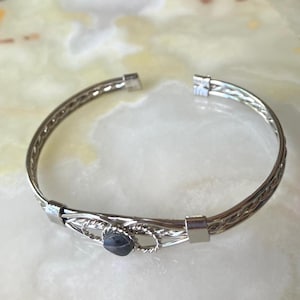 May include: Silver-toned bracelet with a woven design and a small, dark blue gemstone. The bracelet has an open design and is set against a light-colored background. The metal has a polished finish, reflecting light.