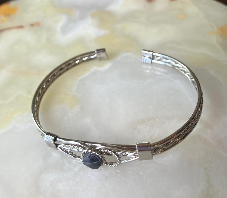 May include: Silver-toned bracelet with a woven design and a small, dark blue gemstone. The bracelet has an open design and is set against a light-colored background. The metal has a polished finish, reflecting light.