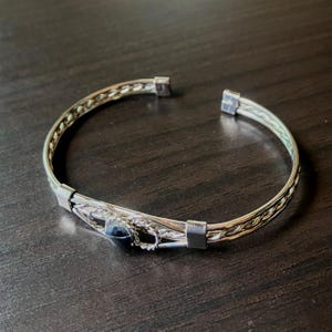 May include: Silver-toned cuff bracelet with a woven design and a dark blue stone. The bracelet has a simple, open design with a decorative center section. The metal has a polished finish, and the stone is round.