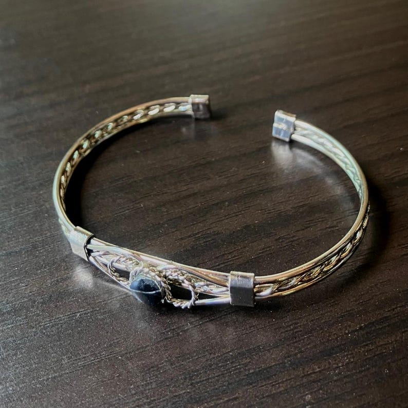 May include: Silver-toned cuff bracelet with a woven design and a dark blue stone. The bracelet has a simple, open design with a decorative center section. The metal has a polished finish, and the stone is round.