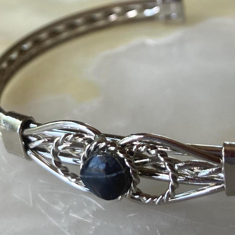 May include: Silver-toned bracelet with a dark blue, faceted bead at the center. The bracelet features a woven design with multiple strands of metal and a clasp closure. The bead is approximately 0.25 inches in diameter.