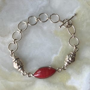 May include: A silver-toned chain bracelet featuring a central marquise-shaped red stone, two textured silver-toned beads, and a toggle clasp closure. The bracelet is constructed with a series of circular links connected to the central focal point.