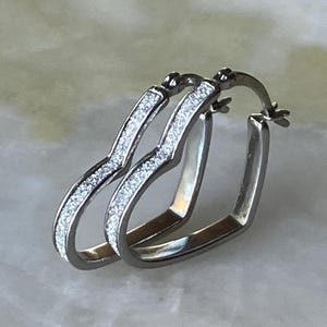 May include: A pair of silver-toned hoop earrings shaped like hearts. The front-facing surface of each heart-shaped hoop is textured with a fine, sparkling glitter finish, while the inner and side edges feature a smooth, polished metallic surface.