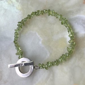 May include: A bracelet composed of small, irregular, light green peridot chip beads strung together. The bracelet features a silver-toned metal toggle clasp consisting of a circular ring and a bar engraved with the word LUCK in capital letters.