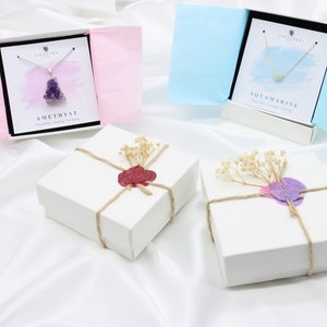 May include: Two white gift boxes tied with twine and sealed with wax, one red and one purple. Two open gift boxes display necklaces, one with a purple amethyst pendant and the other with a blue aquamarine pendant. The boxes are on a white satin surface.