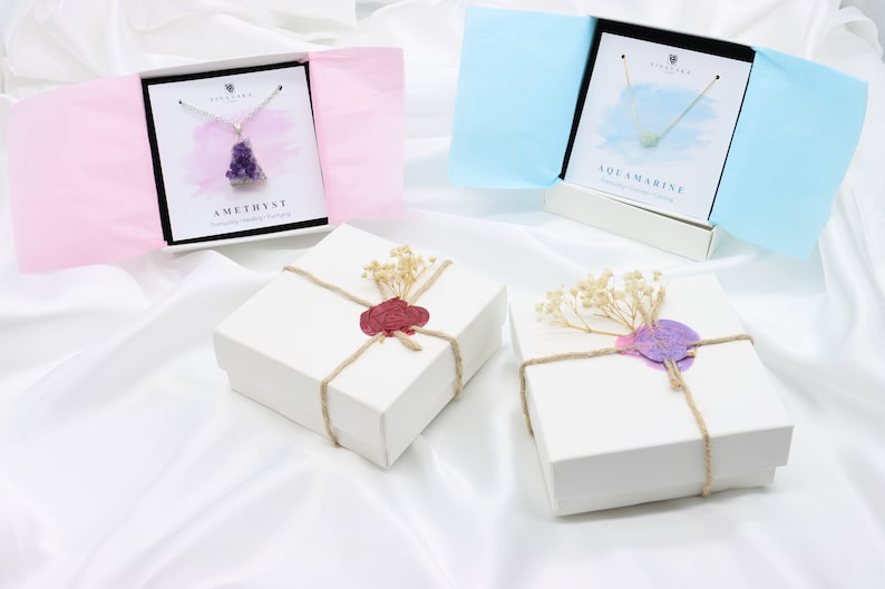 May include: Two white gift boxes tied with twine and sealed with wax, one red and one purple. Two open gift boxes display necklaces, one with a purple amethyst pendant and the other with a blue aquamarine pendant. The boxes are on a white satin surface.